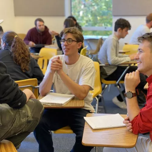 Three students engaged in a discussion in a classroom, with notebooks on their desks.