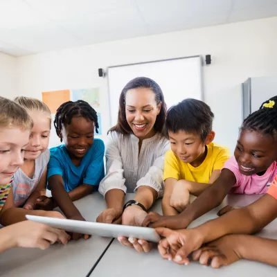 Elementary Students gathered around an elementary school teacher looking at a tablet computer.
