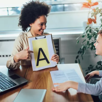 A teacher and a student talking, the teacher is holding up a piece of paper with a large letter A.