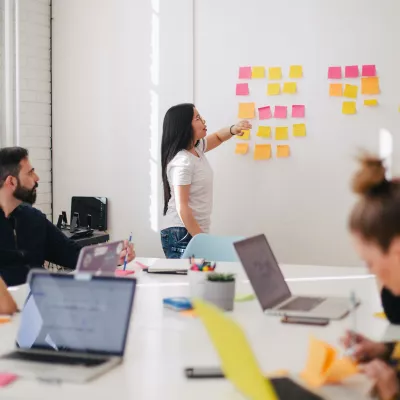 A woman presenting a board full of post it notes to a table full of people
