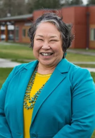 Laural Ballew wearing a teal blazer over a yellow blouse and a green beaded necklace. She is smiling.