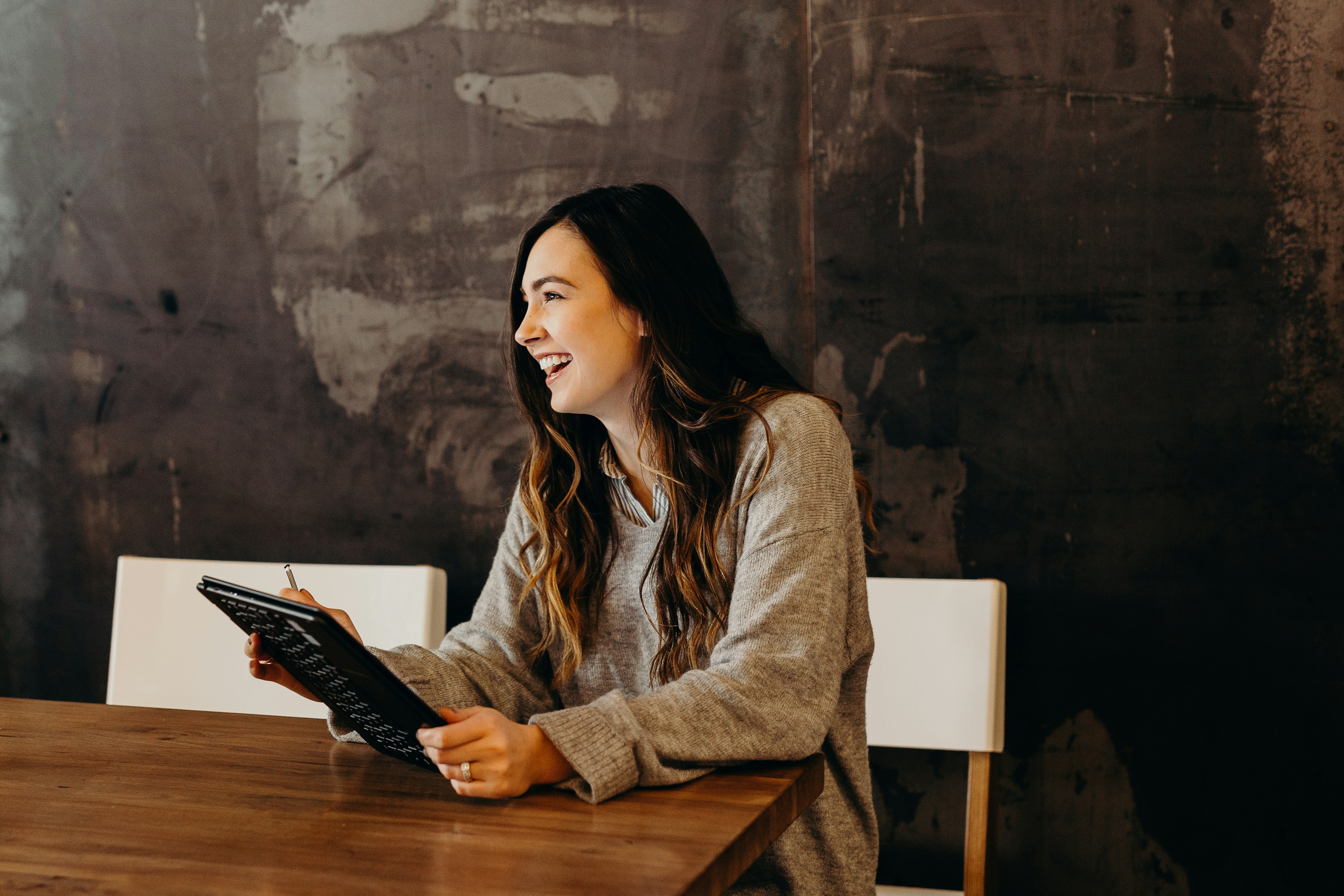 Smiling student in a gray sweater, holding a tablet while sitting in front of a blackboard