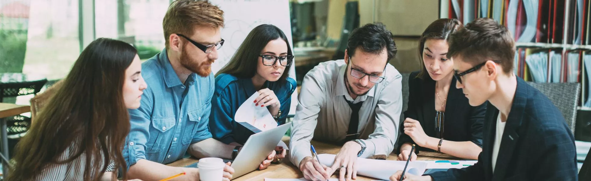 Group of students in the Business Administration degree program sit around a desk, listening to a male student speak