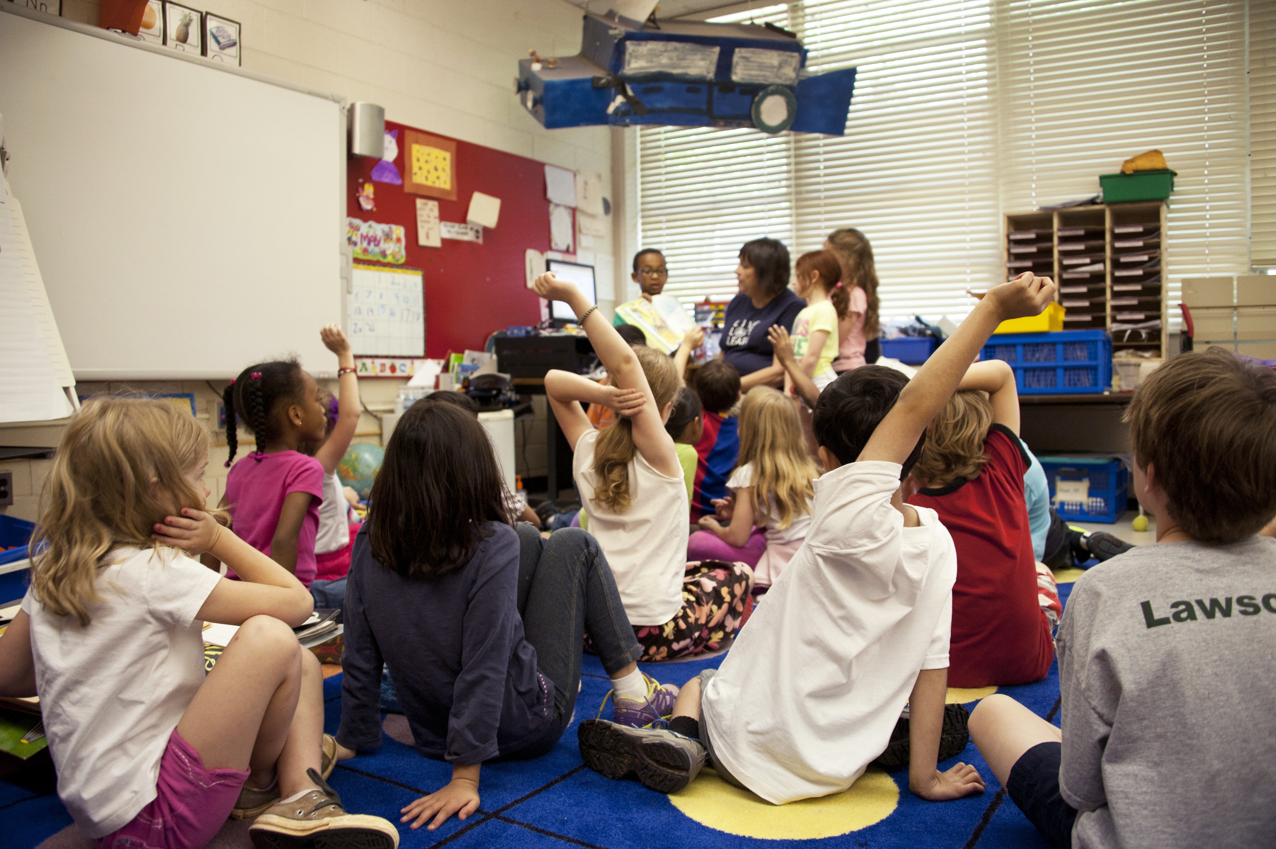 Children gathered on a rug in a classroom, some raising their hands
