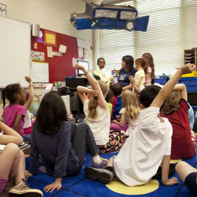 Children gathered on a rug in a classroom, some raising their hands
