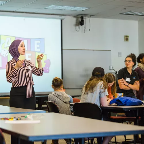 An instructor stands at the projector screen in front of a class of students.