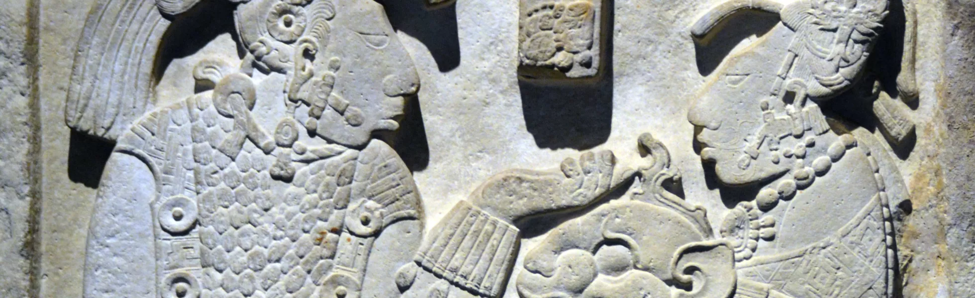 Mayan stone carving of a royal lady giving her king a jaguar mask and shield. They wear elaborate clothing, headdresses, and body jewelry.