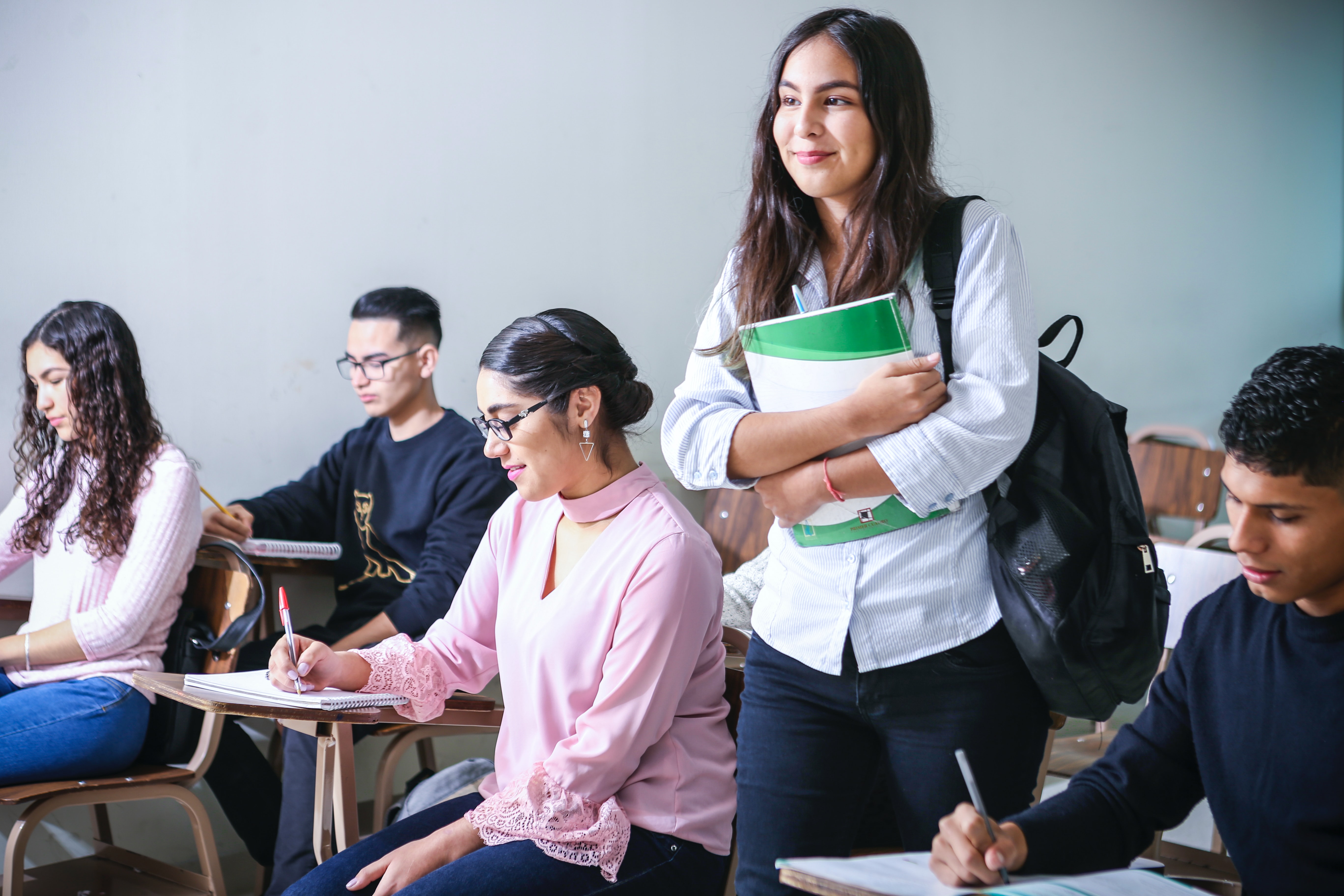 Students in a classroom, most seated at desk with one standing holding a green folder