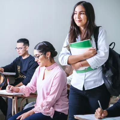 Students in a classroom, most seated at desk with one standing holding a green folder