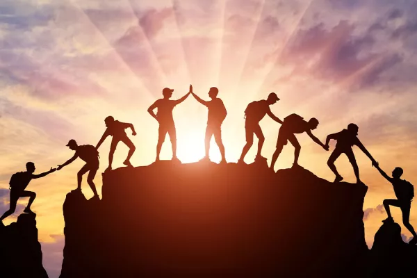 Silhouettes of people helping each other climb and reach the summit against a vibrant sunset.