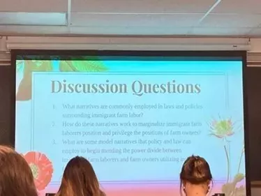 A projector screen displays "Discussion Questions" about narratives, power, and labor, likely for a class or meeting. Three people's backs are visible in the foreground.
