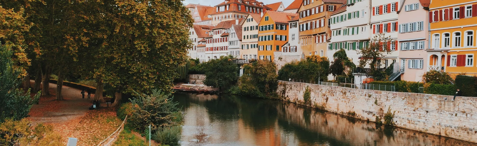 Timber-framed German houses along a river canal. On the other side of the river is a park with large trees and a walking path.