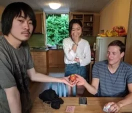 3 students standing and sitting around a table, an apple is passed between 2 of them.