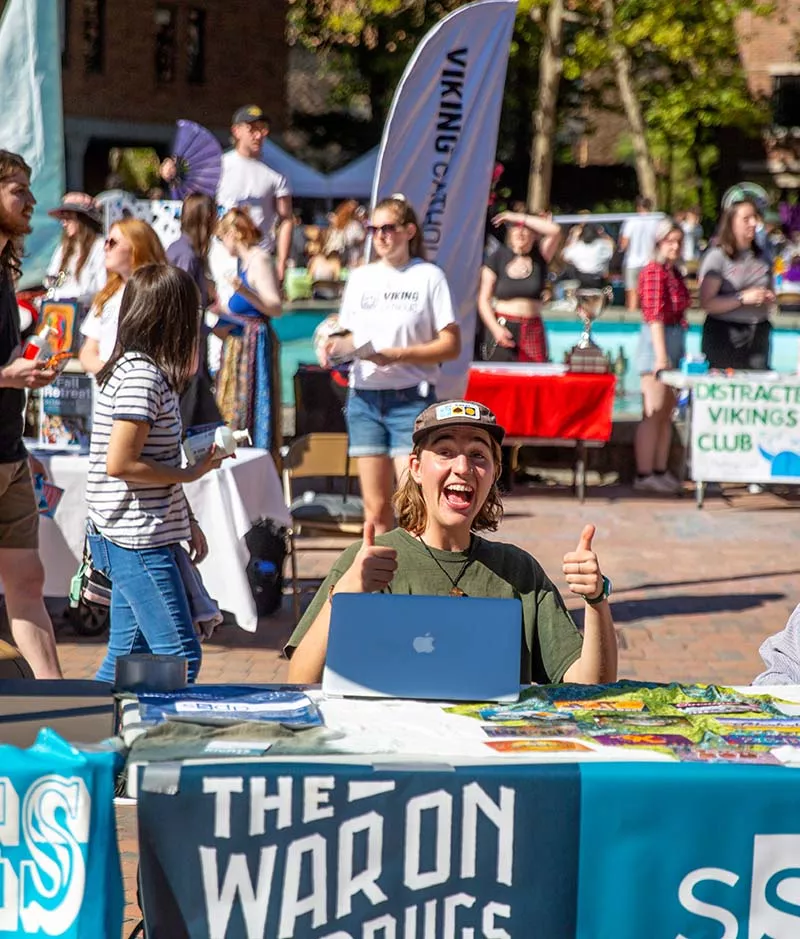 A student sitting at a table at the info fair, giving an enthusiastic double thumbs up