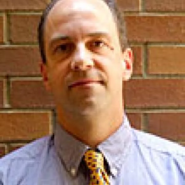 John Thompson stands in front of a brick wall in a button down shirt and tie.