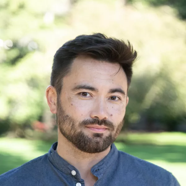 Short dark brown hair and a clean brown beard. Wearing a buttoned blue shirt. 