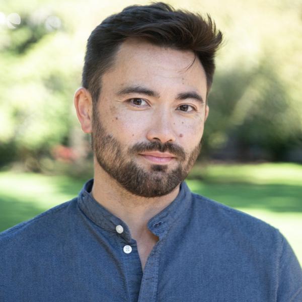 Short dark brown hair and a clean brown beard. Wearing a buttoned blue shirt. 