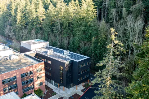 Aerial view of a modern, dark-wood building with solar panels on its roof, nestled in a lush evergreen forest. Adjacent brick building visible.