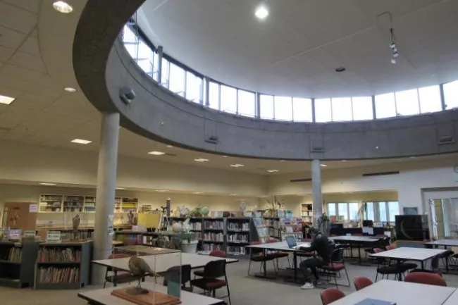 A modern library interior with circular skylight, tables, chairs, and shelves filled with books.