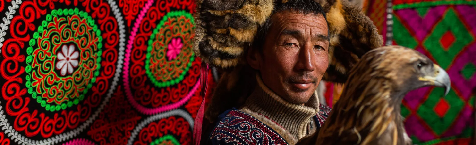 A Mongolian man standing in a bright yurt with a golden eagle on his arm. He wears a fur hat. Medals are hung on the richly embroidered walls of the yurt.