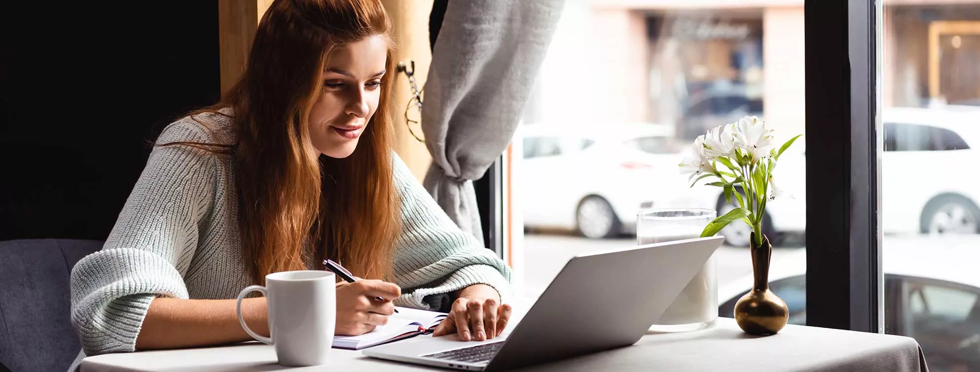 A student sits at a coffee shop, in front of an open window, with a cup of coffee beside them.