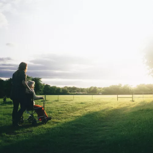 A person pushes another person in a wheelchair across a sunny grass field.