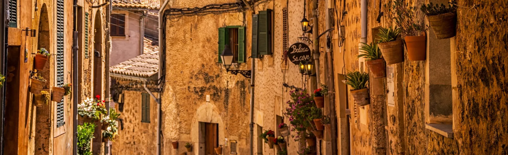 A warm and sunny cobblestone street lined with stone and plaster buildings with green shutters and the small plant pots suspended on the walls.