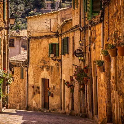 A warm and sunny cobblestone street lined with stone and plaster buildings with green shutters and the small plant pots suspended on the walls.