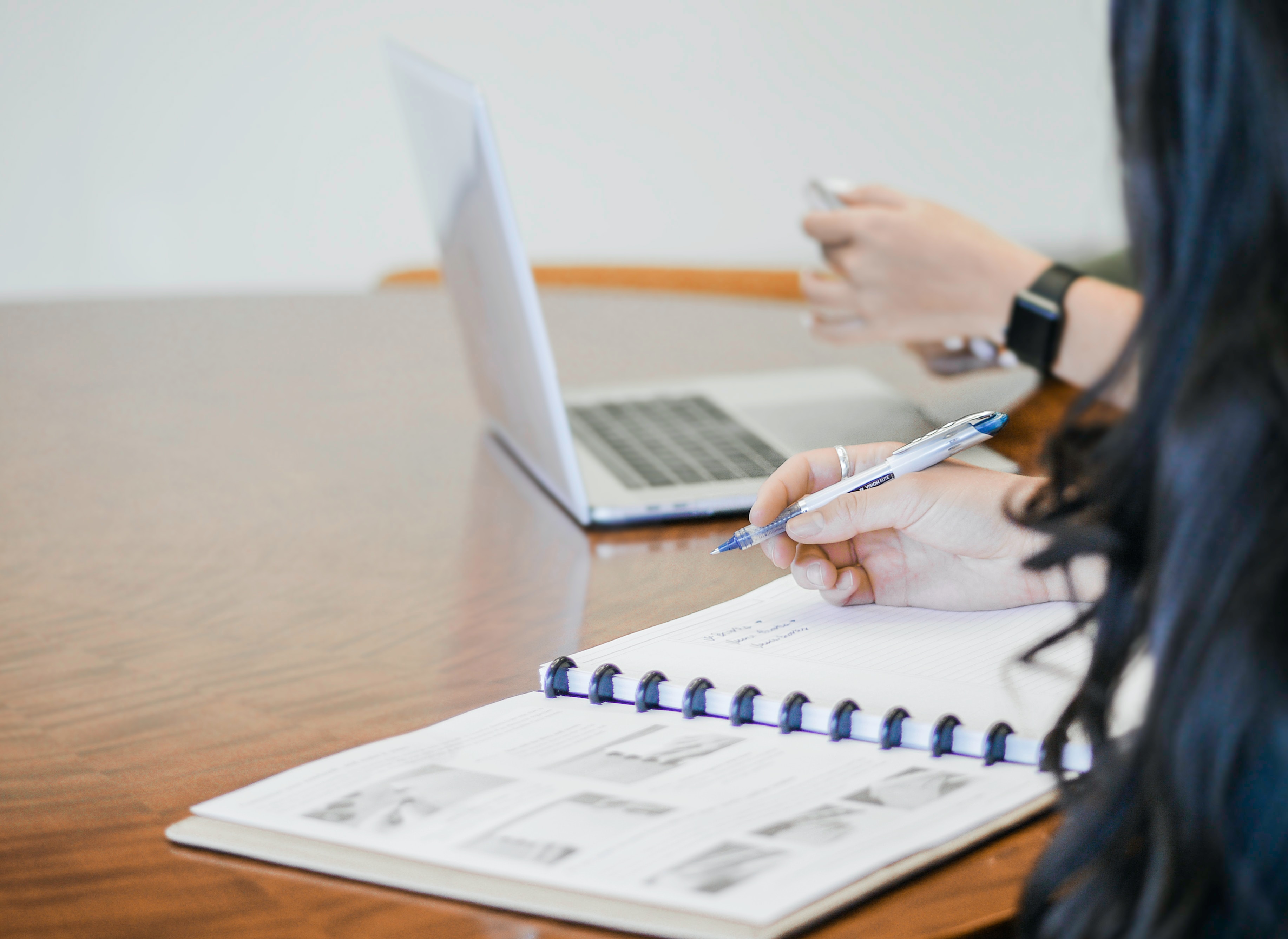 A person working on a spiral notebook and laptop on a desk