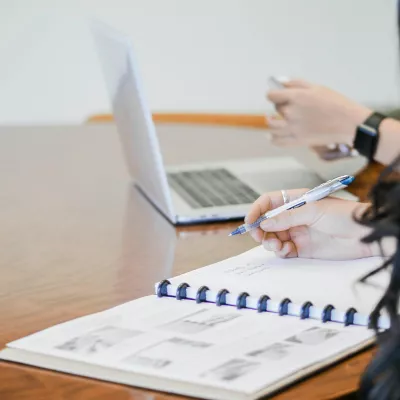 A person working on a spiral notebook and laptop on a desk