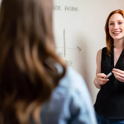 A teacher at a whiteboard in conversation with a person whose back is to the camera