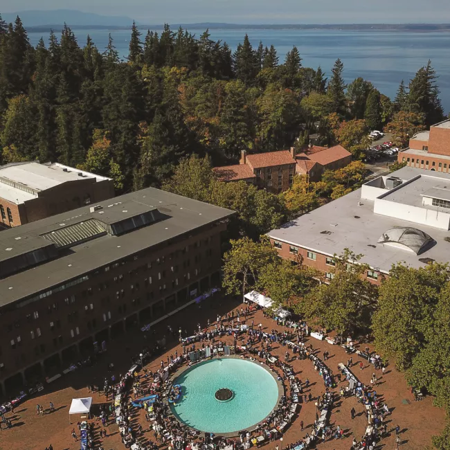 Aerial view of a bustling university courtyard with a fountain, surrounded by trees and overlooking a large body of water.