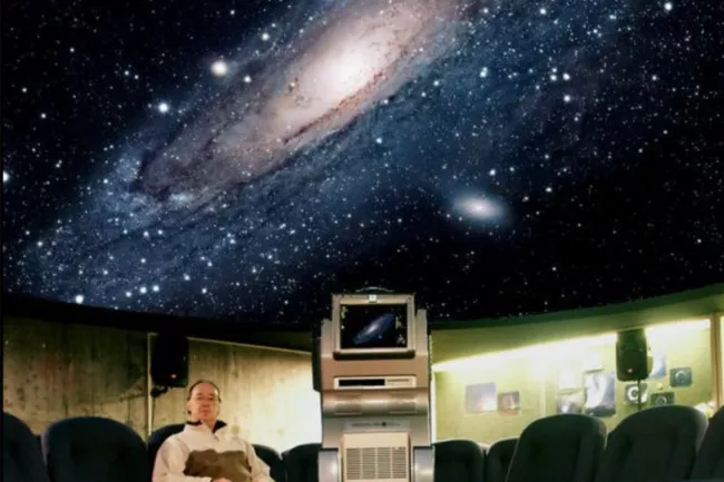Interior of a planetarium with a projection of a galaxy on the domed ceiling and a person seated among rows of chairs.