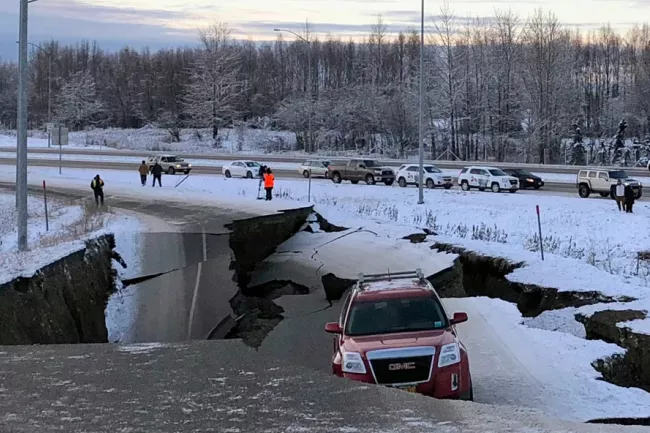 A large crack splits a snow-covered road, near several cars and onlookers.  A red SUV is partially positioned over the fissure. Winter landscape.